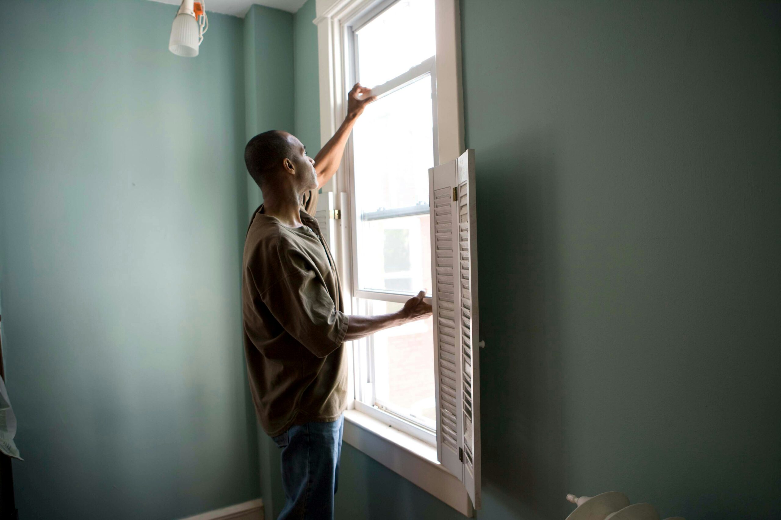 A person standing in a room with green walls opens a white window, letting in natural light while practicing mosquito control. One hand is on the window, and the other holds the latch. A white shutter is partially open below the window.