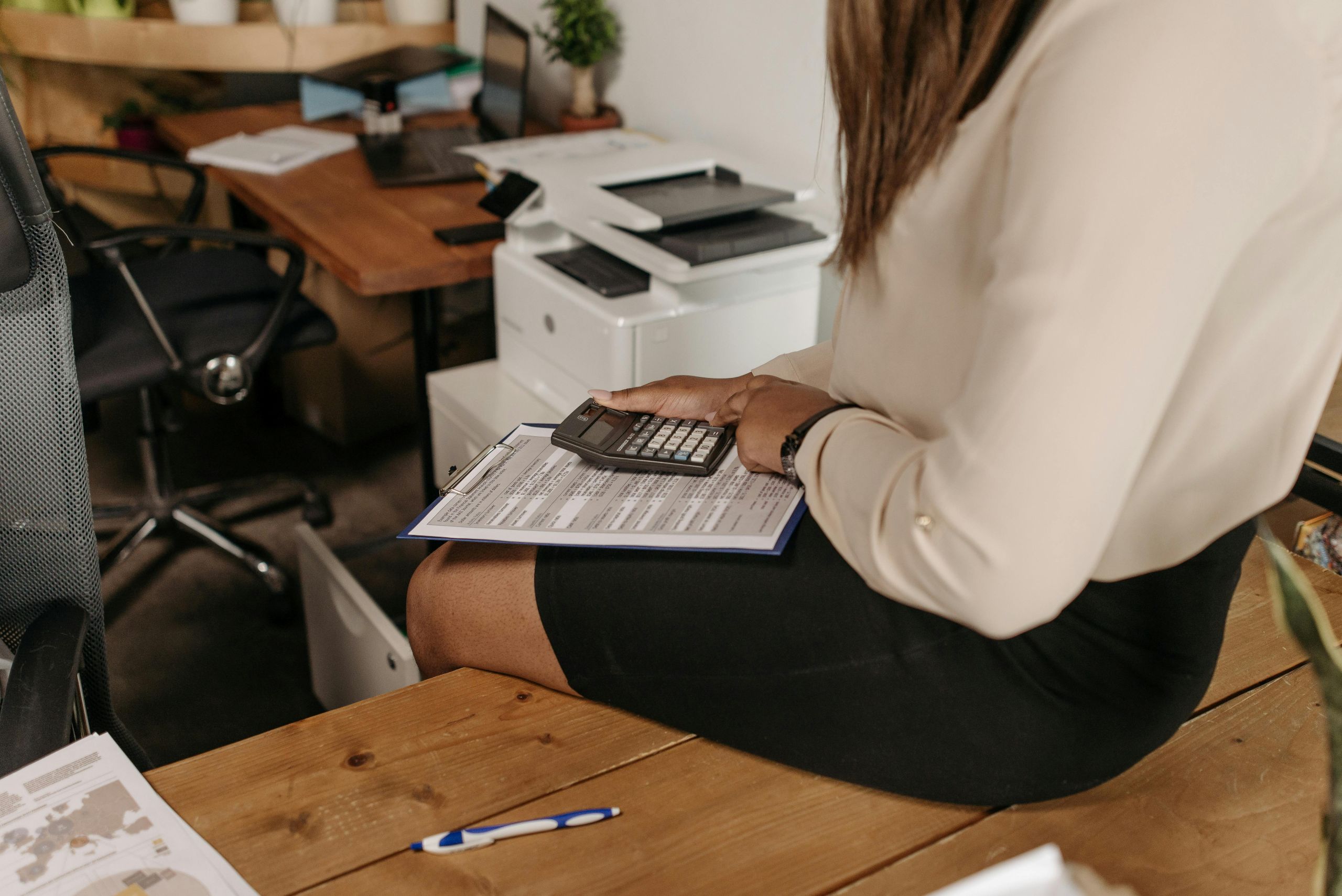 A woman in business attire sits on a desk, holding a clipboard and using a calculator. Office equipment, papers, and a pen are visible on surrounding desks in the commercial pest control workspace.