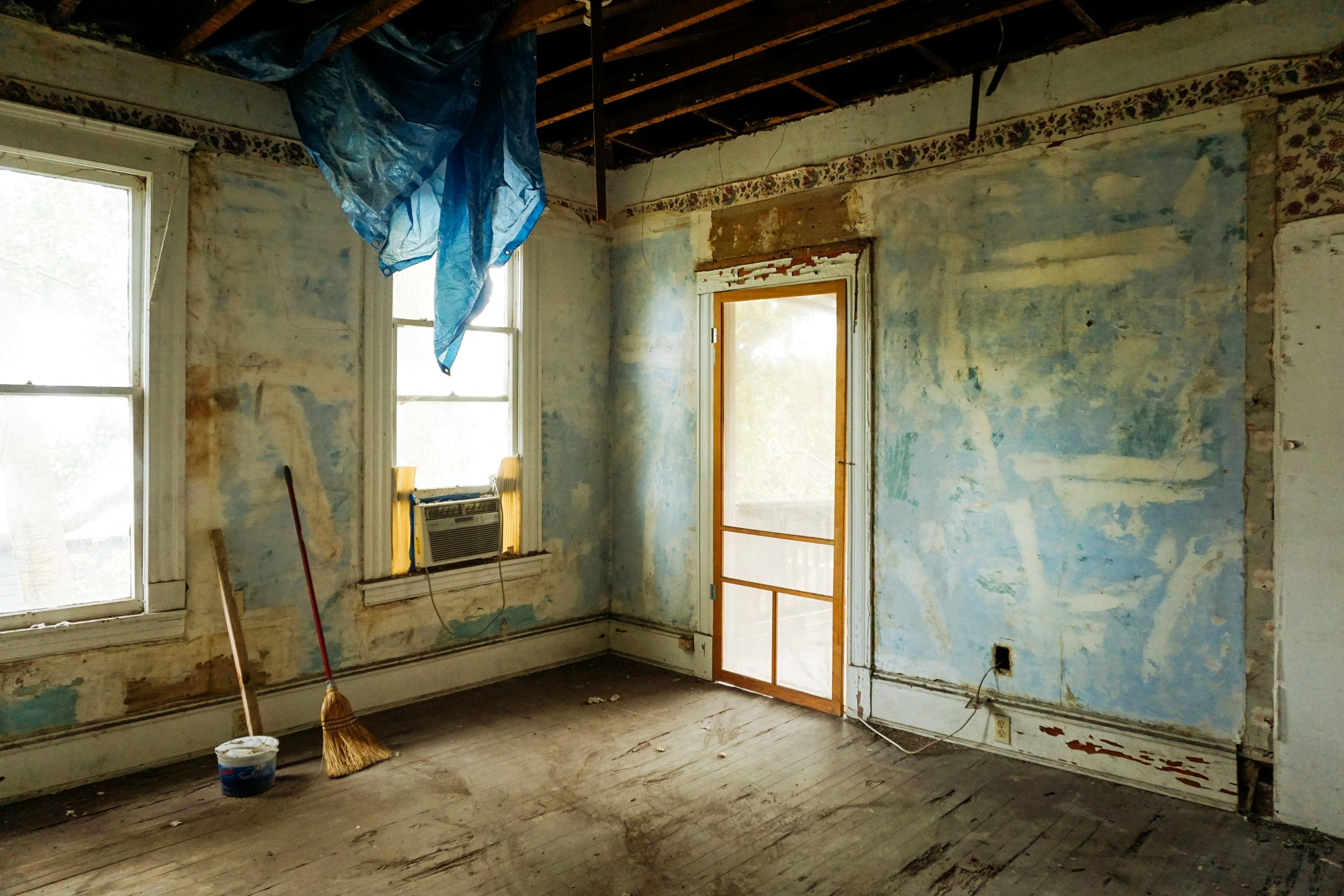 A rundown room with peeling blue paint, exposed ceiling beams, and blue tarp hints at common entry points pests use during winter, as cracks around two windows or the old air conditioner may let insects inside. A broom and bucket rest on the worn wooden floor.