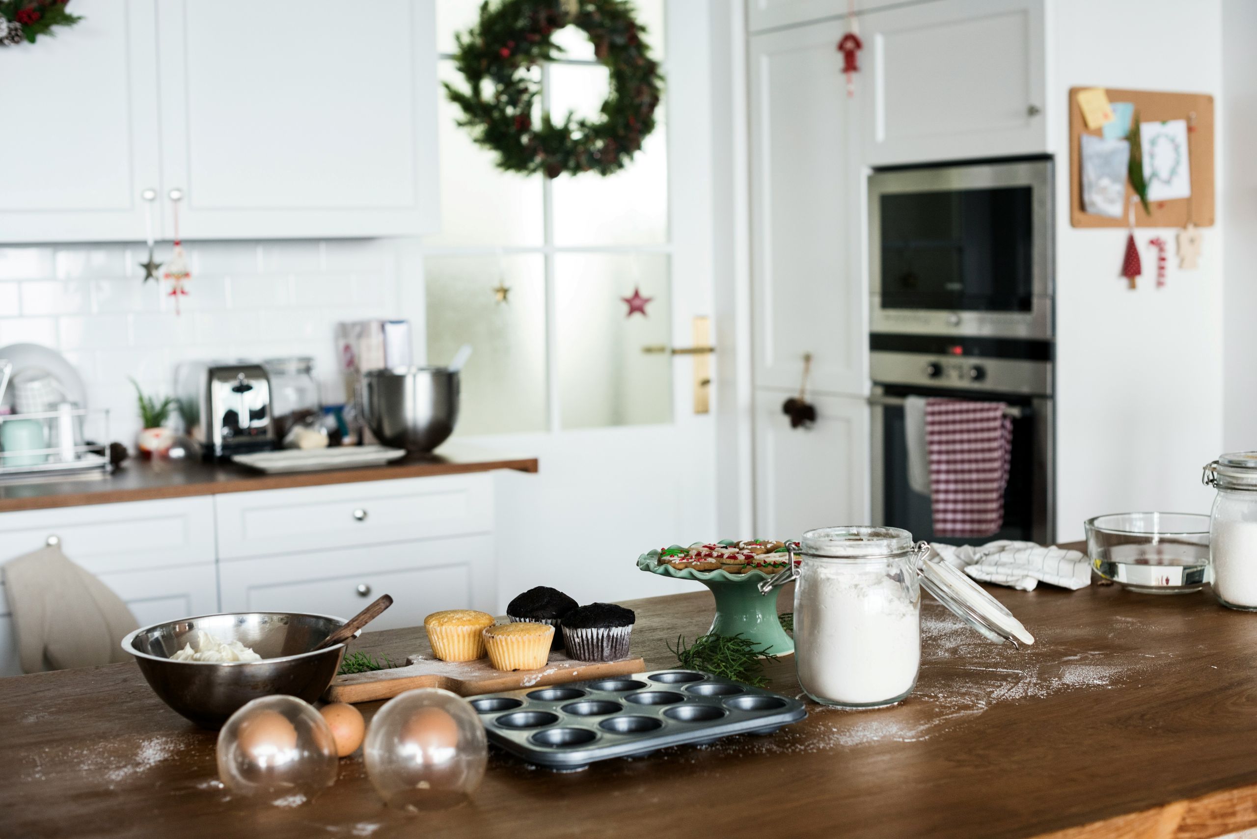 A kitchen with baking supplies on a wooden counter, including flour, cupcakes, mixing bowls, and utensils. Christmas wreaths and decorations hang in the background, creating a festive atmosphere free from pests.