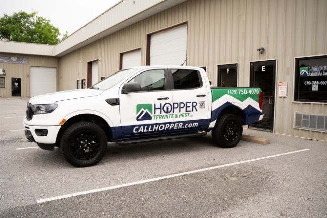 White pickup truck with Hopper Termite & Pest branding parked outside a beige industrial building; the truck displays a phone number and website for pest control services.