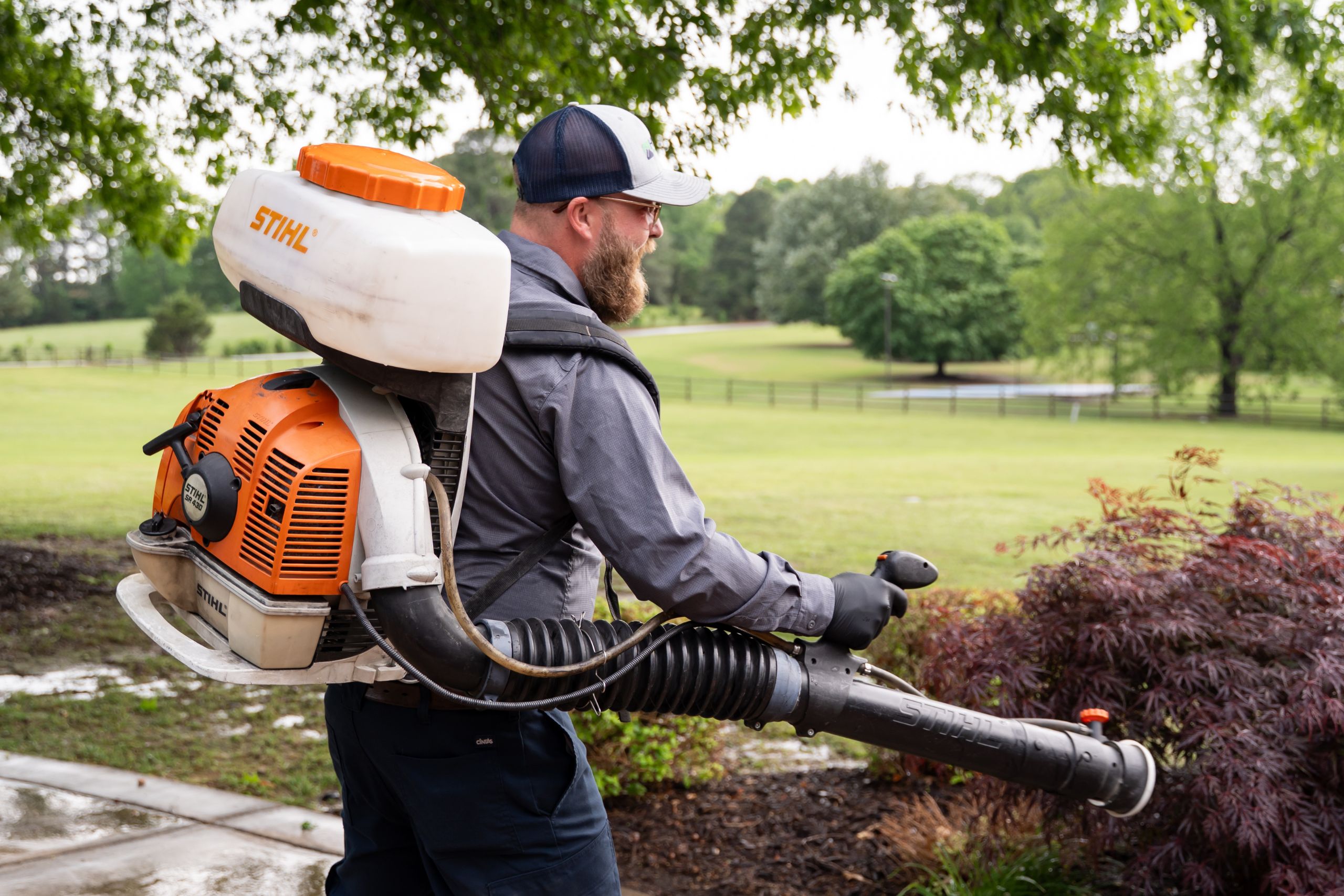 Mosquito Control Service in NWA and Mountain Home A man wearing a cap and gloves uses a STIHL leaf blower in a park-like setting with green grass, trees, and a cloudy sky in the background.