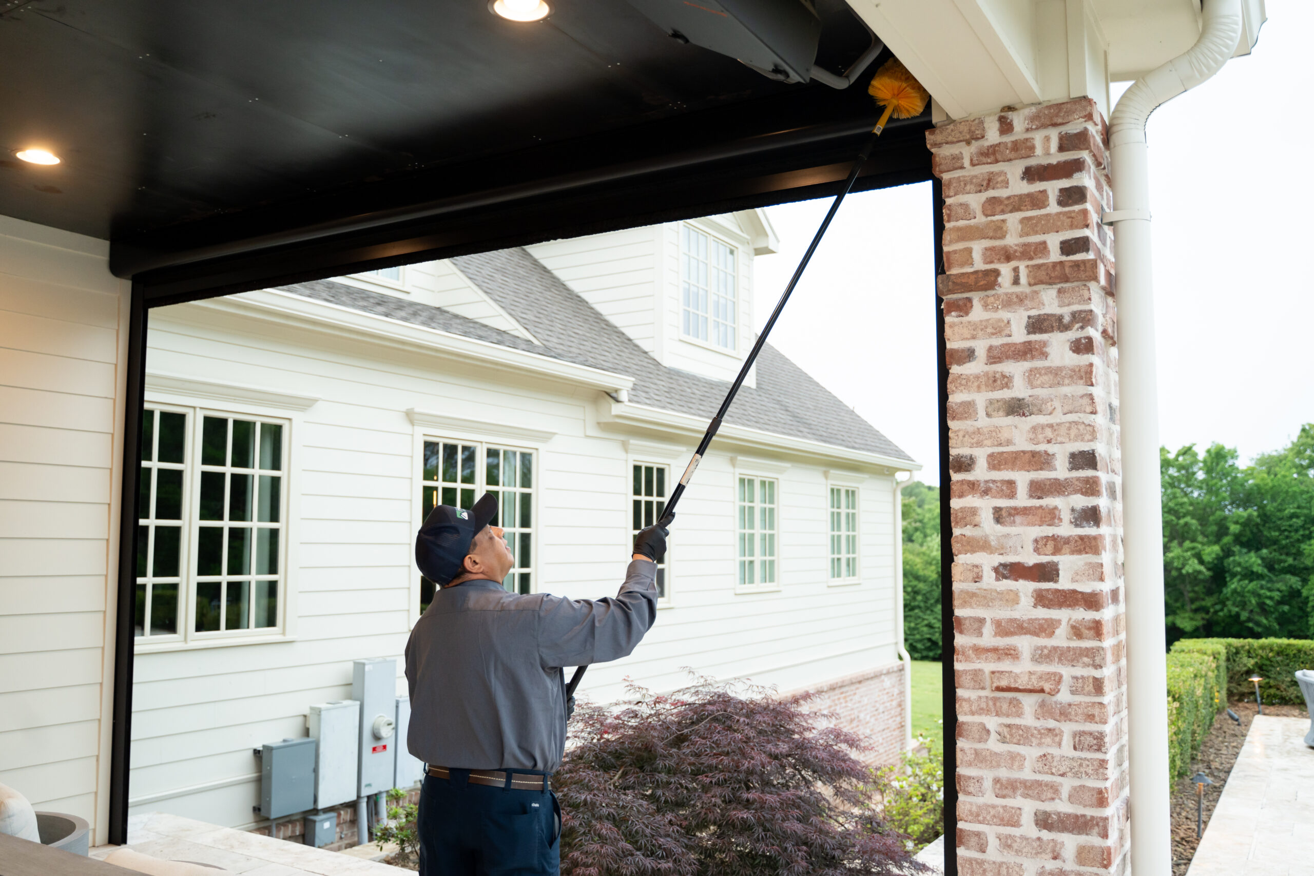 Spider control in NWA and Mountain Home A person wearing a cap and uniform uses a long-handled tool to clean the upper corner of a patio area near a brick column, with a house and greenery visible in the background.