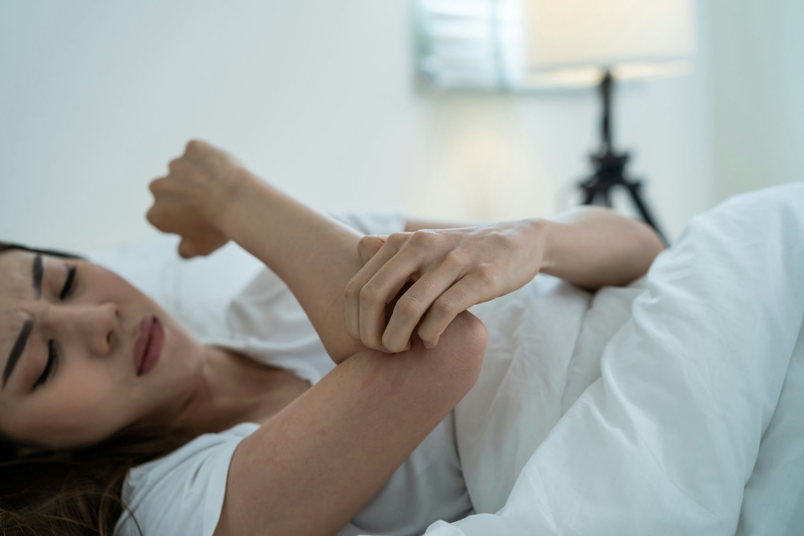 A young woman lies in bed, scratching her arm with an expression of discomfort.